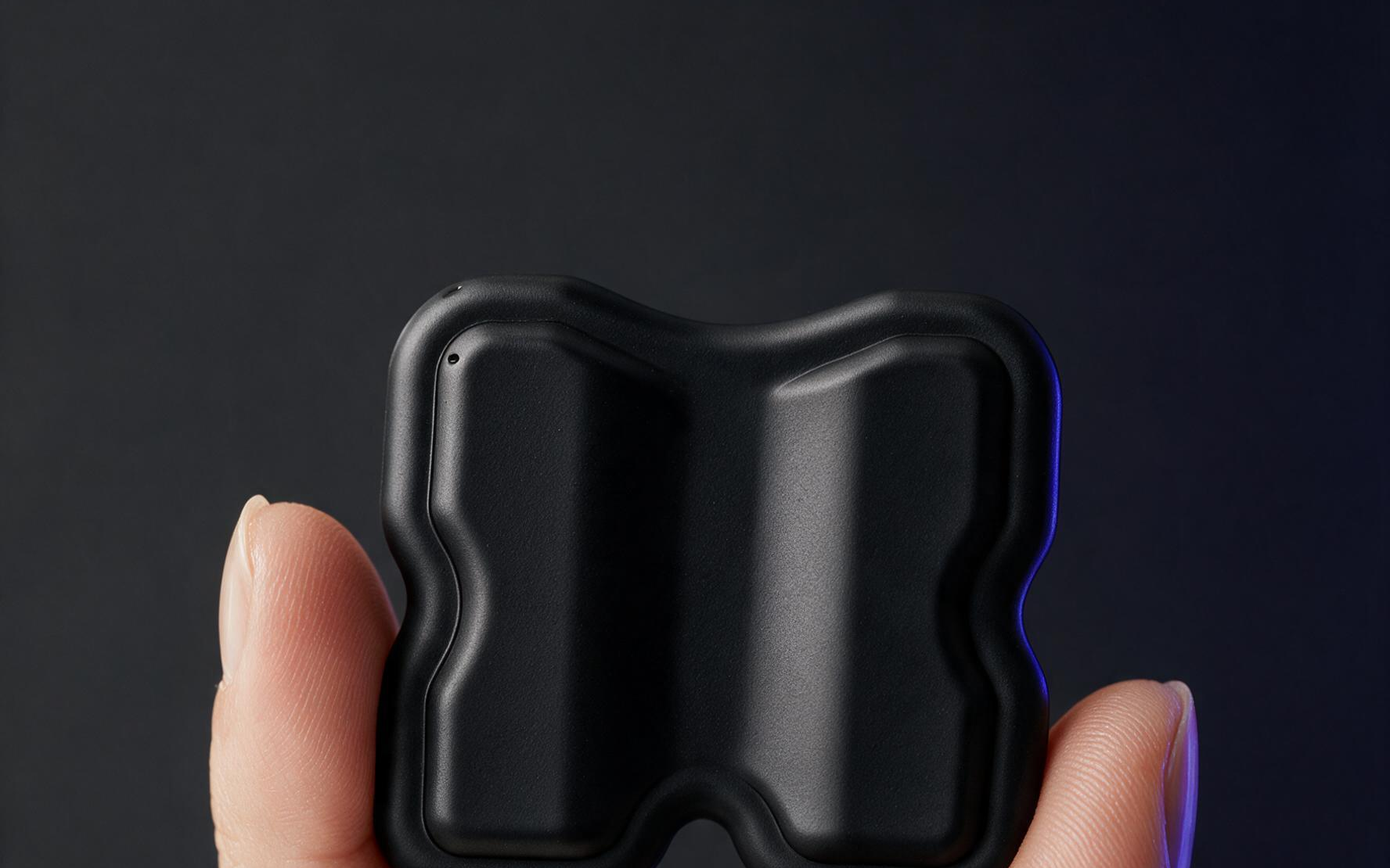 Fingers holding the KTRL patch: H-shaped black conductive silicone single-use patch pinched between thumb and forefinger